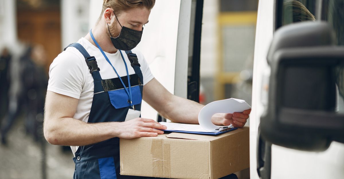 A delivery worker wearing a face mask checks a package next to a van, ensuring safety measures.